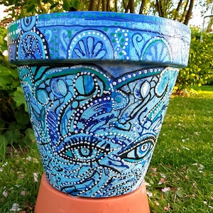 Può includere: Vaso di fiori in ceramica dipinto a mano con un vivace design blu e turchese. Il vaso presenta motivi intricati, tra cui occhi stilizzati e motivi floreali, con accenti a pois bianchi. Poggia su una base in terracotta.