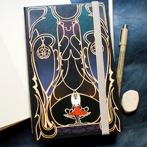 Puede incluir: Un diario negro y dorado con un diseño decorativo y una ilustración de personaje. El diario está junto a un cuaderno abierto, un lápiz, un bolígrafo y una goma de borrar. Los objetos están sobre una superficie azul oscuro.