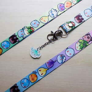 Puede incluir: Dos cordones coloridos con una variedad de personajes de slime de dibujos animados. Los cordones son verde y azul, y morado y azul. Un llavero plateado con un amuleto de slime de dibujos animados está unido a los cordones.