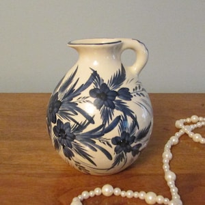 Op de afbeelding: Een kleine witte keramische kan met een blauw bloemenmotief en een handvat. Een witte parelketting met een gouden kwast ligt op een houten oppervlak.