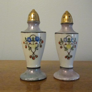 May include: A pair of vintage salt and pepper shakers with a floral design. The shakers are white with a light purple glaze and gold accents. The floral design features red, yellow, and blue flowers.