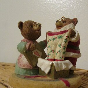 Puede incluir: Una figura navide&ntilde;a de dos osos marrones. Un oso lleva un traje de Pap&aacute; Noel y sostiene un pijama de estampado verde y blanco. El otro oso lleva un vestido azul y rosa y sostiene una caja de regalo.
