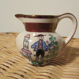 May include: A small white ceramic creamer with a brown rim and a painted scene of a person in a blue shirt and a red hat standing in front of a building. The creamer has a brown handle.