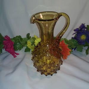 May include: Amber-colored glass pitcher with a textured, hobnail design. The pitcher has a curved handle and a flared spout. The glass is a warm, golden hue, and the hobnail pattern covers the bulbous body. Decorative flowers in shades of pink, orange, yellow, and purple are in the background.