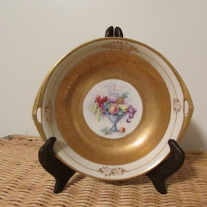 May include: A decorative porcelain dish with a gold interior and rim, featuring a central fruit arrangement. The white border has floral accents. The fruit arrangement includes grapes, peaches, and other fruits. The dish has a diameter of approximately 20 cm.