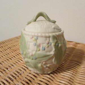 May include: A white ceramic sugar bowl with a green lid and floral design. The lid has a handle and the bowl is decorated with green leaves and pink and blue flowers.