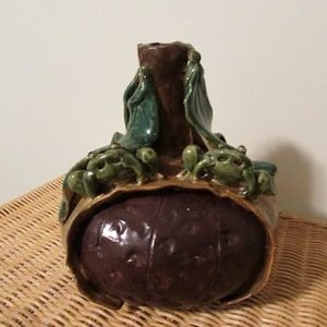 May include: A ceramic vase with a brown, green, and purple glaze. The vase is decorated with four green frogs and has a round, bulbous body.