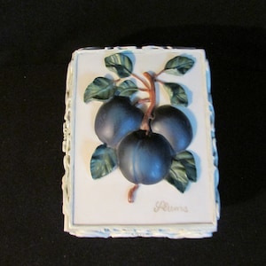 May include: A white ceramic box with a raised image of three plums on a branch with green leaves. The box has a white border with a decorative design. The word "Plums" is written in cursive on the bottom right corner of the box.