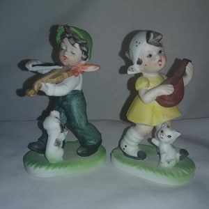 May include: A pair of porcelain figurines of a boy and girl playing music. The boy is wearing a green hat and playing a violin. The girl is wearing a yellow dress and playing a mandolin. Both figurines have a white dog or cat beside them.
