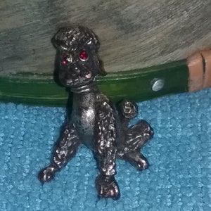 May include: A small, silver-colored poodle figurine with red rhinestone eyes. The poodle is sitting with its legs outstretched and its tail curled.