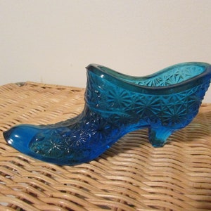 May include: A vintage, turquoise blue glass shoe-shaped decorative piece. The shoe features a detailed starburst pattern and a small heel. The open top suggests it could be used as a small vase or trinket holder.