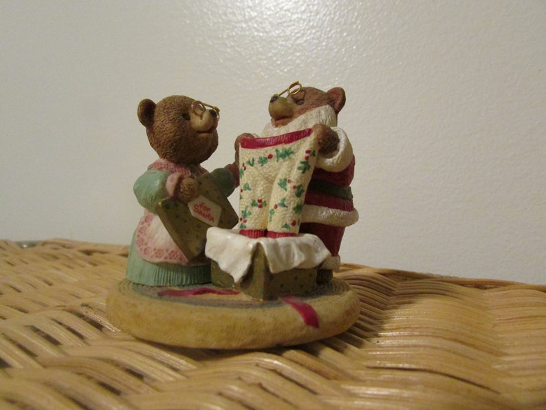 Puede incluir: Una figura navide&ntilde;a de dos osos marrones. Un oso lleva un traje de Pap&aacute; Noel y sostiene un pijama de estampado verde y blanco. El otro oso lleva un vestido azul y rosa y sostiene una caja de regalo.