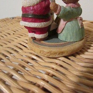 Puede incluir: Una peque&ntilde;a figura de cer&aacute;mica de Santa Claus y una mujer sosteniendo un rollo. La figura est&aacute; sobre una base multicolor con las palabras "By Joelle" escritas en la parte inferior.