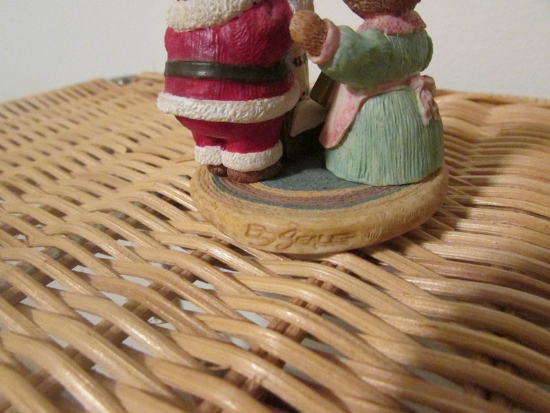 Puede incluir: Una peque&ntilde;a figura de cer&aacute;mica de Santa Claus y una mujer sosteniendo un rollo. La figura est&aacute; sobre una base multicolor con las palabras "By Joelle" escritas en la parte inferior.