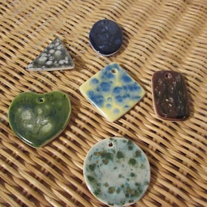 May include: A collection of handmade ceramic pendants in various shapes: a triangle, oval, diamond, rectangle, heart, and circle. Each pendant features unique glaze patterns in shades of green, blue, brown, and white. The pendants have a hole for stringing.