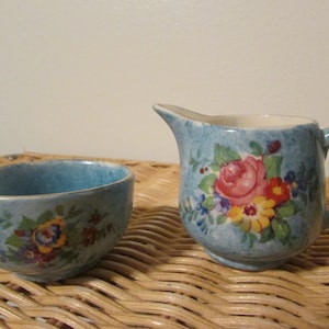 May include: A small, light blue creamer pitcher and matching bowl. Both pieces are decorated with a floral pattern featuring pink, red, yellow, and blue flowers. The creamer has a handle and spout.