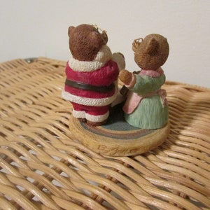 Puede incluir: Una figura navide&ntilde;a de dos osos vestidos como Pap&aacute; Noel y una mujer. El oso vestido como Pap&aacute; Noel lleva un traje rojo y blanco con un cintur&oacute;n negro. El otro oso lleva un vestido verde con un delantal rosa. La figura est&aacute; sobre una base redonda con un patr&oacute;n marr&oacute;n y blanco.