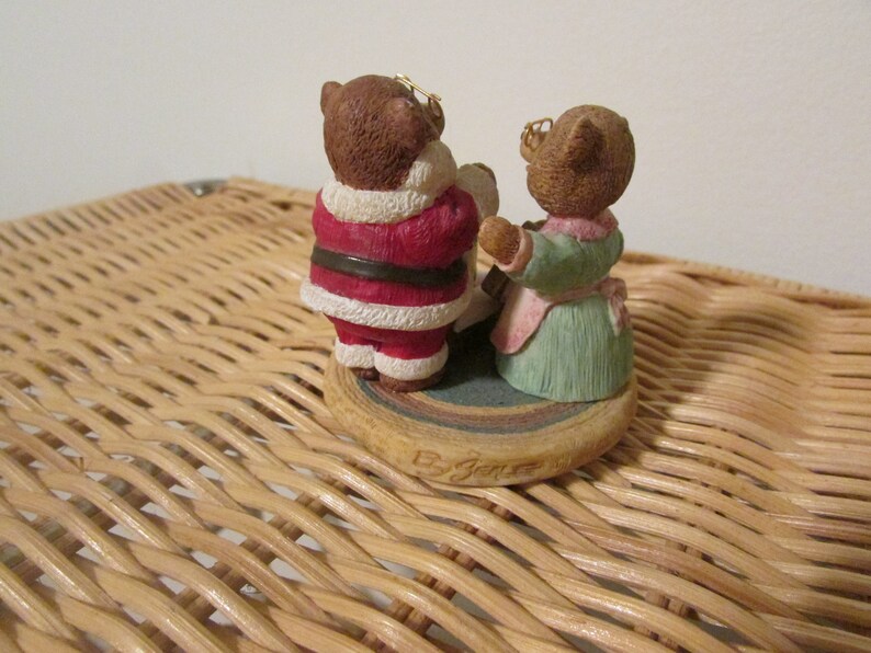 Puede incluir: Una figura navide&ntilde;a de dos osos vestidos como Pap&aacute; Noel y una mujer. El oso vestido como Pap&aacute; Noel lleva un traje rojo y blanco con un cintur&oacute;n negro. El otro oso lleva un vestido verde con un delantal rosa. La figura est&aacute; sobre una base redonda con un patr&oacute;n marr&oacute;n y blanco.