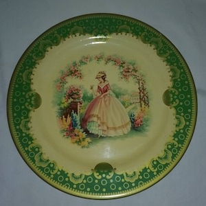 May include: A vintage tin plate with a green and gold border. The center features a woman in a white dress standing in a garden with flowers.