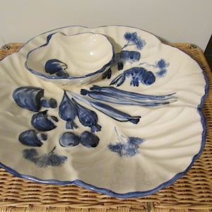 Op de afbeelding: Een witte keramische serveerschaal met een geschulpte rand en een kleine, bevestigde kom. De schaal is versierd met blauw geschilderde groenten en zeevruchten. De rand van de schaal en de kom zijn blauw.