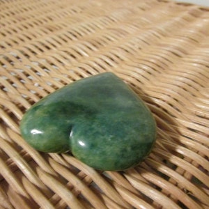 May include: A heart-shaped green stone with a mottled surface. The stone is resting on a woven wicker surface.