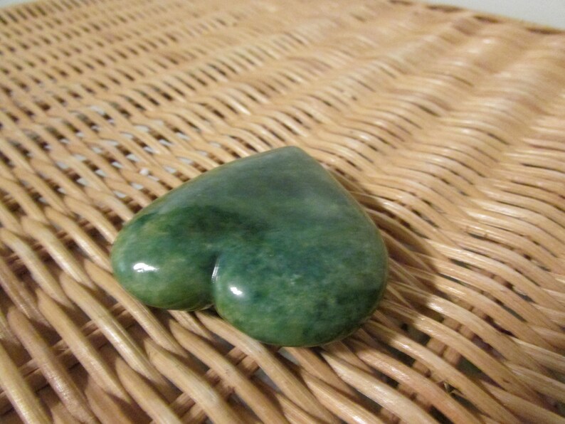 May include: A heart-shaped green stone with a mottled surface. The stone is resting on a woven wicker surface.