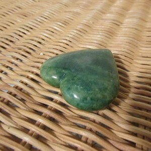 May include: A heart-shaped green stone with a smooth, polished surface. The stone is resting on a woven wicker surface.