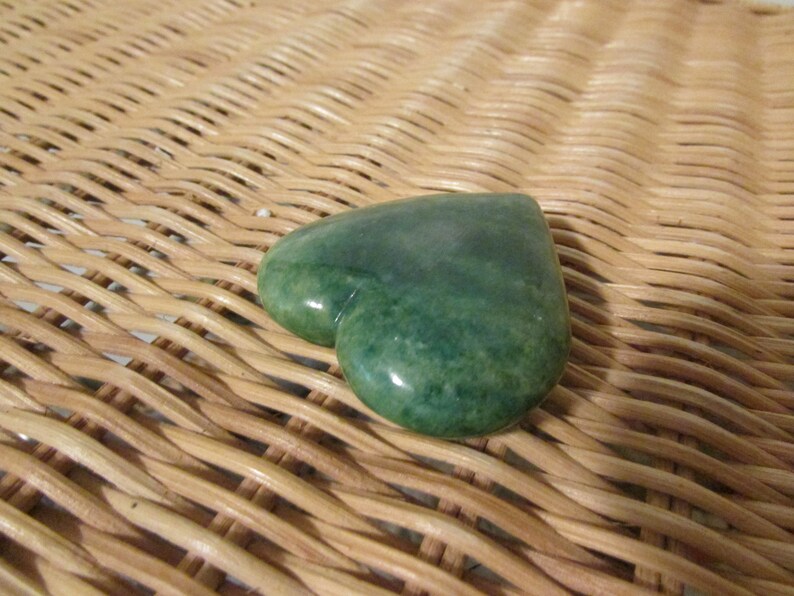 May include: A heart-shaped green stone with a smooth, polished surface. The stone is resting on a woven wicker surface.