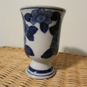 Puede incluir: Un jarrón de cerámica blanca con un diseño floral azul. El jarrón tiene una base de pedestal.