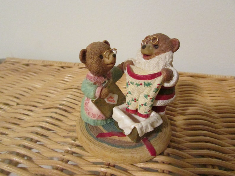 Puede incluir: Dos osos de peluche marrones que llevan gafas. Un oso sostiene un su&eacute;ter a rayas rojo y verde con hojas de acebo y bayas. El otro oso sostiene un sobre peque&ntilde;o con las palabras "Para Santa" escritas en &eacute;l. Los osos est&aacute;n de pie sobre una base redonda de madera con una cinta roja.