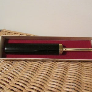 May include: A black and gold cigarette lighter in a white box with a red interior. The lighter has a long, thin design and a gold tip.