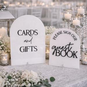 May include: Two arched white acrylic signs with black lettering. The larger sign reads CARDS and GIFTS. The smaller sign reads PLEASE SIGN OUR guest BOOK. Both signs feature a small white circular base and are displayed on a table.
