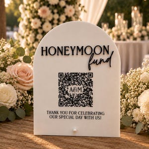 May include: A white, arched sign with the words "HONEYMOON fund" in black script. It includes a QR code and the text "THANK YOU FOR CELEBRATING OUR SPECIAL DAY WITH US!" A pearl is at the bottom.