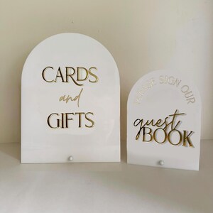 May include: Two white arched acrylic signs with gold lettering. The larger sign reads CARDS and GIFTS in a mix of block and script fonts. The smaller sign reads PLEASE SIGN OUR guest BOOK in a mix of block and script fonts. Both signs have a small peg.