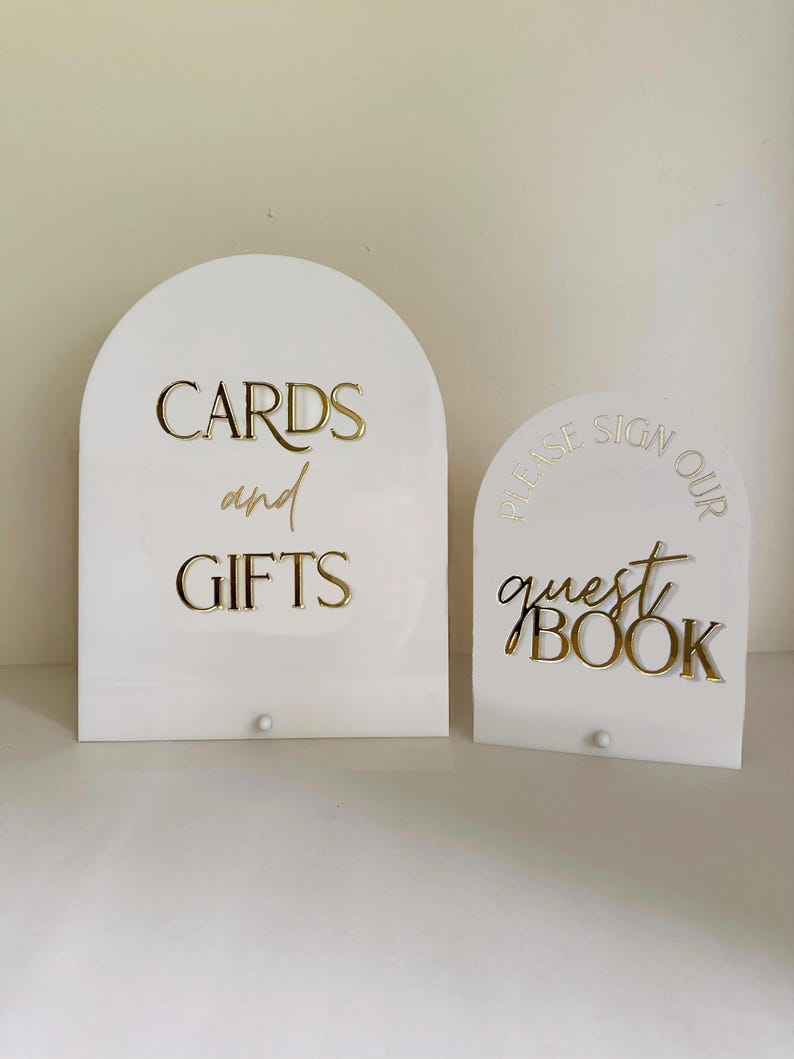 May include: Two white arched acrylic signs with gold lettering. The larger sign reads CARDS and GIFTS in a mix of block and script fonts. The smaller sign reads PLEASE SIGN OUR guest BOOK in a mix of block and script fonts. Both signs have a small peg.