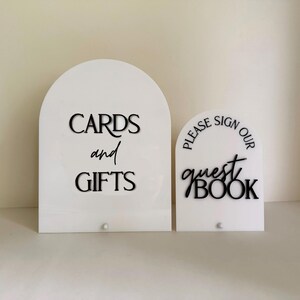 May include: Two white arched acrylic signs with black lettering. The larger sign reads CARDS and GIFTS in a mix of serif and script fonts. The smaller sign reads PLEASE SIGN OUR guest BOOK in a mix of serif and script fonts. Both have a small stud.