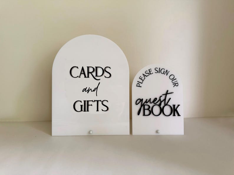 May include: Two white arched acrylic signs with black lettering. The larger sign reads CARDS and GIFTS in a mix of serif and script fonts. The smaller sign reads PLEASE SIGN OUR guest BOOK in a mix of serif and script fonts. Both have a small stud.