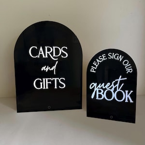 May include: Two arched black acrylic signs with white lettering. The larger sign reads CARDS and GIFTS. The smaller sign reads PLEASE SIGN OUR guest BOOK. Both signs feature a smooth, reflective surface and a rounded top edge design.