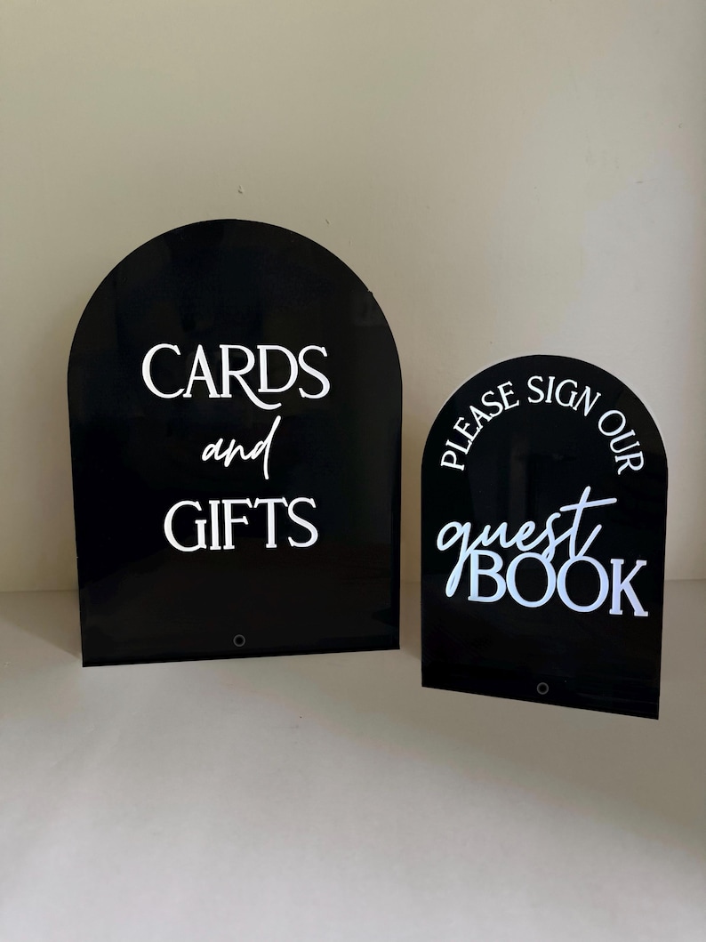 May include: Two arched black acrylic signs with white lettering. The larger sign reads CARDS and GIFTS. The smaller sign reads PLEASE SIGN OUR guest BOOK. Both signs feature a smooth, reflective surface and a rounded top edge design.