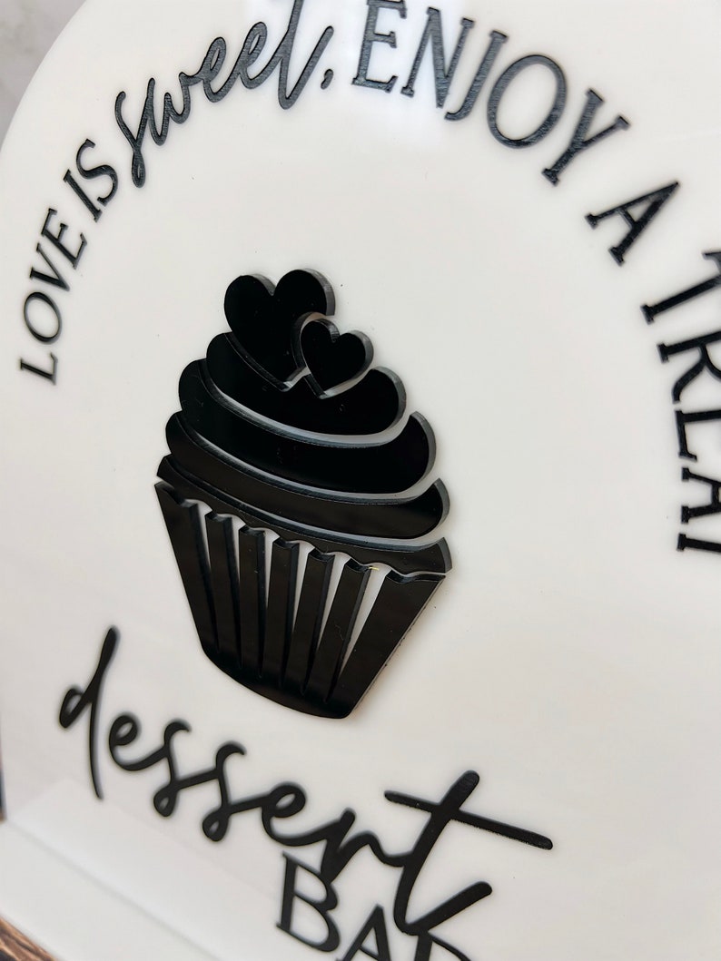 May include: White sign with black text and a black cupcake graphic. The text reads "Love is sweet. Enjoy a little dessert bar."