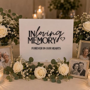 May include: A white rectangular memorial sign with black lettering that reads IN loving MEMORY and FOREVER IN OUR HEARTS, accompanied by a small heart icon. The sign is surrounded by white roses, greenery, framed photographs, and lit candles.