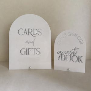 May include: Two arched white acrylic signs with silver lettering. The larger sign reads CARDS and GIFTS, while the smaller sign reads PLEASE SIGN OUR guest BOOK. Each sign features a small circular base attachment at the bottom centre.