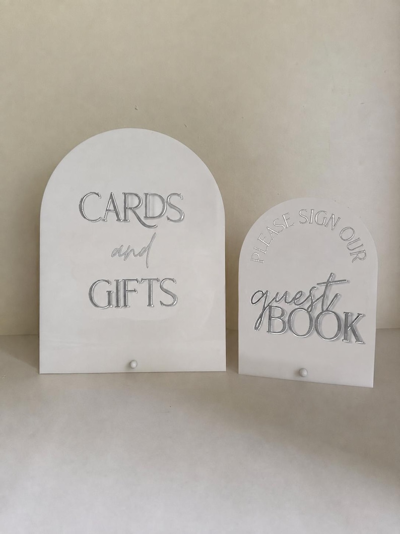 May include: Two arched white acrylic signs with silver lettering. The larger sign reads CARDS and GIFTS, while the smaller sign reads PLEASE SIGN OUR guest BOOK. Each sign features a small circular base attachment at the bottom centre.
