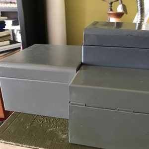 May include: Two gray metal boxes with hinged lids. The boxes are stacked on top of each other. The top box is slightly larger than the bottom box.