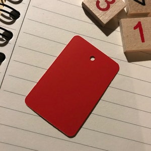 May include: A red rectangular tag with a hole in the top. The tag is on a white lined notebook with wooden blocks with the numbers 1, 3, and 4.