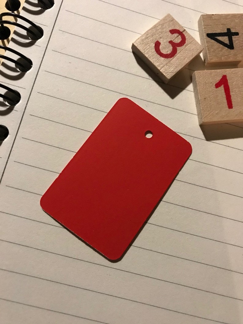 May include: A red rectangular tag with a hole in the top. The tag is on a white lined notebook with wooden blocks with the numbers 1, 3, and 4.