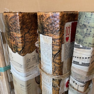 May include: Four vintage-style journals with brown paper covers, each decorated with different paper scraps, including a map, sheet music, and a playing card. The journals are tied with twine and have a rustic, handmade aesthetic.