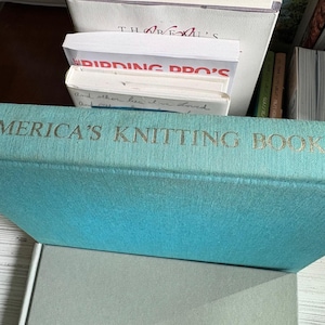 May include: A teal-colored book titled "America's Knitting Book" with gold lettering. The book is positioned in front of other books and papers. The book's spine is visible, showcasing its title. The book appears to be vintage.