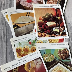 May include: A collection of vintage recipe cards featuring various food and drink ideas. The cards are colorful and feature illustrations of the dishes. Some of the recipes include "Hot Fruits", "Fruit-Cheese Tray with Anise Toast", "Adventure in Space", "Beef Paprika", "Entertaining on a Shoestring", and "Spanish Pizza for the Gang".