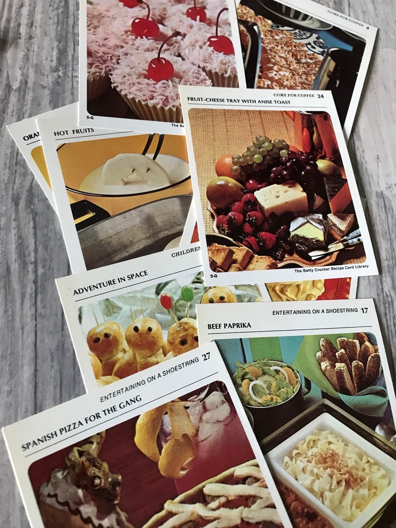May include: A collection of vintage recipe cards featuring various food and drink ideas. The cards are colorful and feature illustrations of the dishes. Some of the recipes include "Hot Fruits", "Fruit-Cheese Tray with Anise Toast", "Adventure in Space", "Beef Paprika", "Entertaining on a Shoestring", and "Spanish Pizza for the Gang".